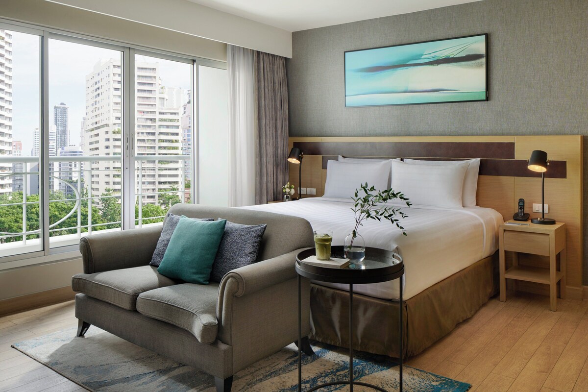 Habitación de hotel con una cama grande, un sofá, una mesita de noche y vistas de la ciudad a través de amplias ventanas.