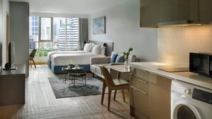 A modern hotel room with a large bed, a dining area, a kitchen with a washing machine, and a view of the cityscape.