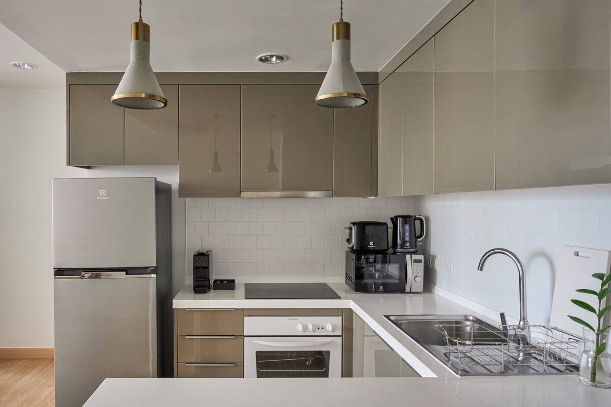 Une cuisine moderne dotée d’appareils électroménagers en acier inoxydable, d’un plan de travail blanc et de luminaires suspendus.