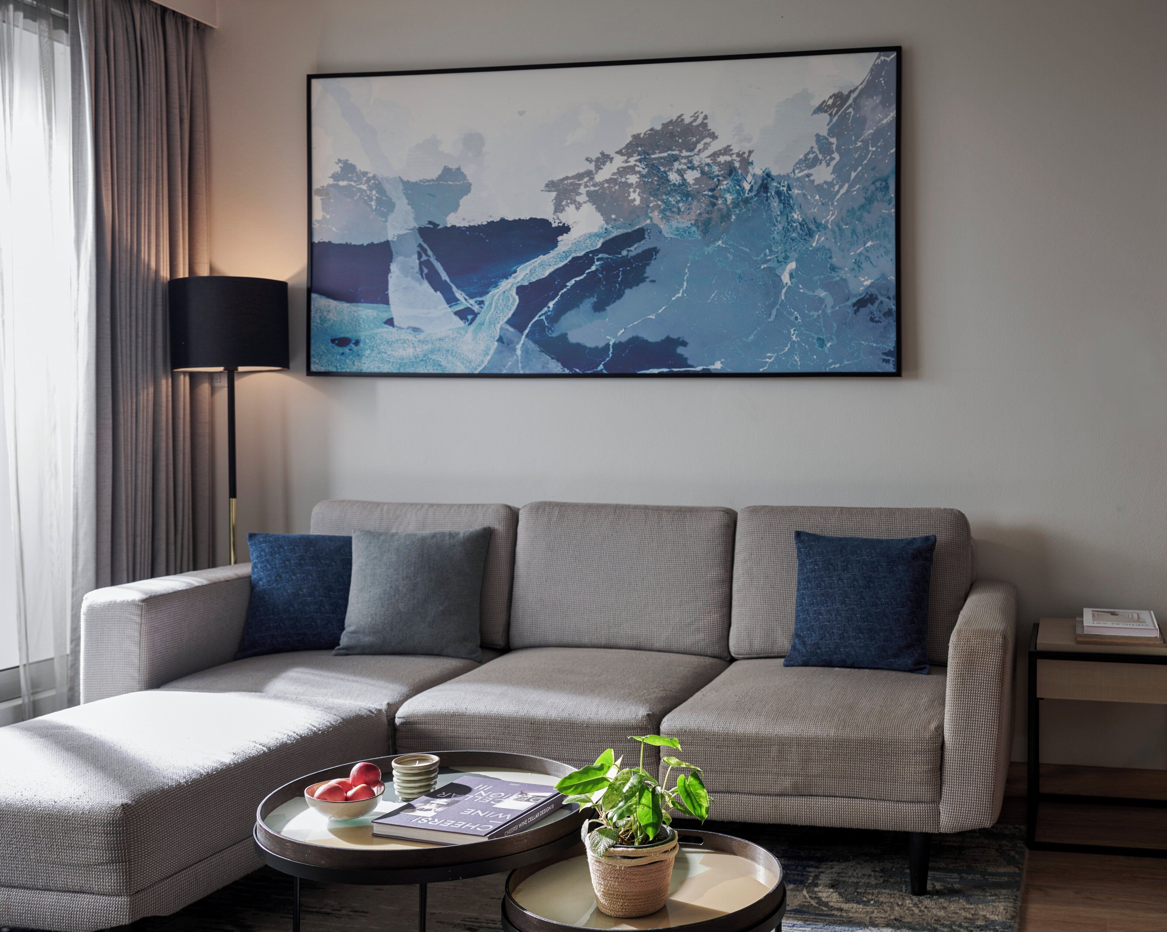 A modern living room with a grey sectional sofa, a coffee table with a book and a plant, and a large abstract painting on the wall.