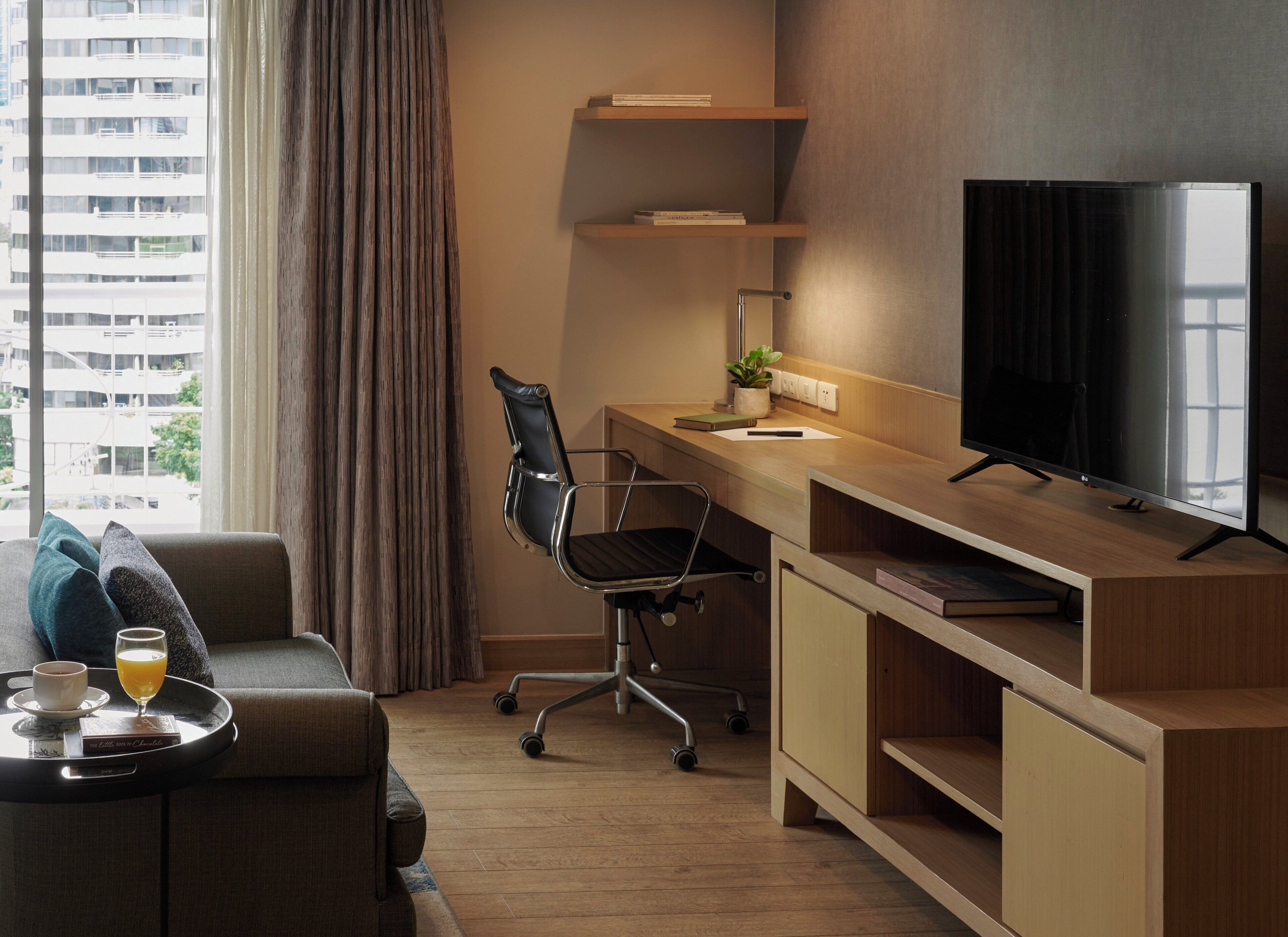 A modern hotel room with a wooden desk, a black office chair, a television, a sofa, and a window with curtains.
