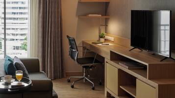 A modern hotel room with a wooden desk, a black office chair, a television, a sofa, and a window with curtains.