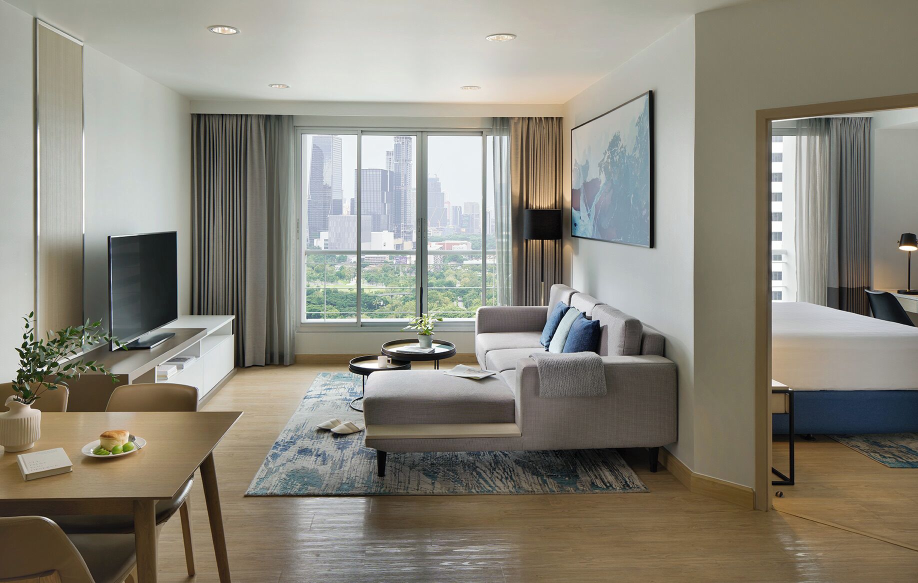 A modern hotel room with a city view, a flat-screen TV, a sofa, a dining area, and a bed.