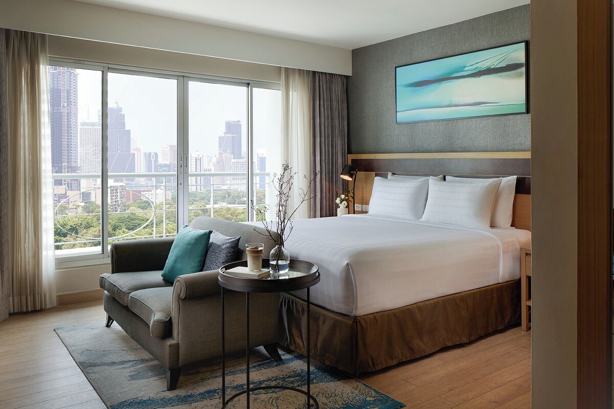 Habitación de hotel con una cama grande, un sofá, una pequeña mesa con una bebida y vistas a la ciudad.