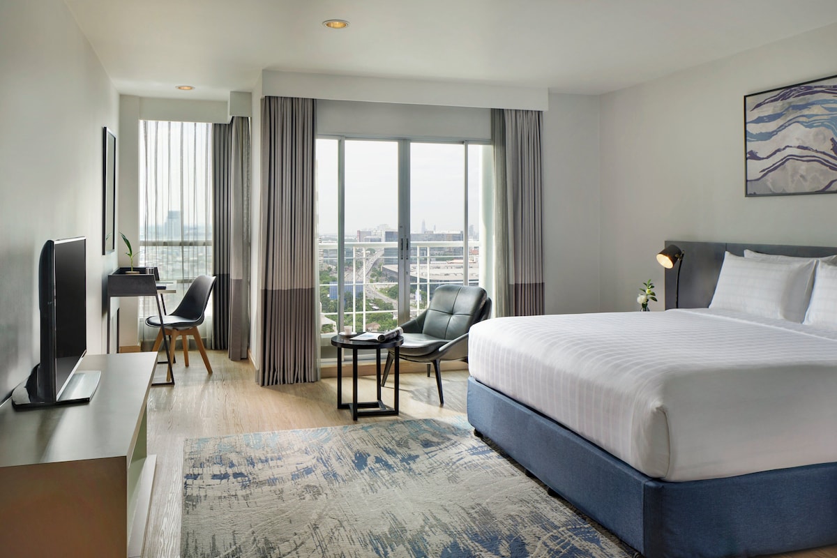 Quarto de hotel com uma cama grande, uma mesa com computador, uma cadeira e vista da cidade através de amplas janelas.