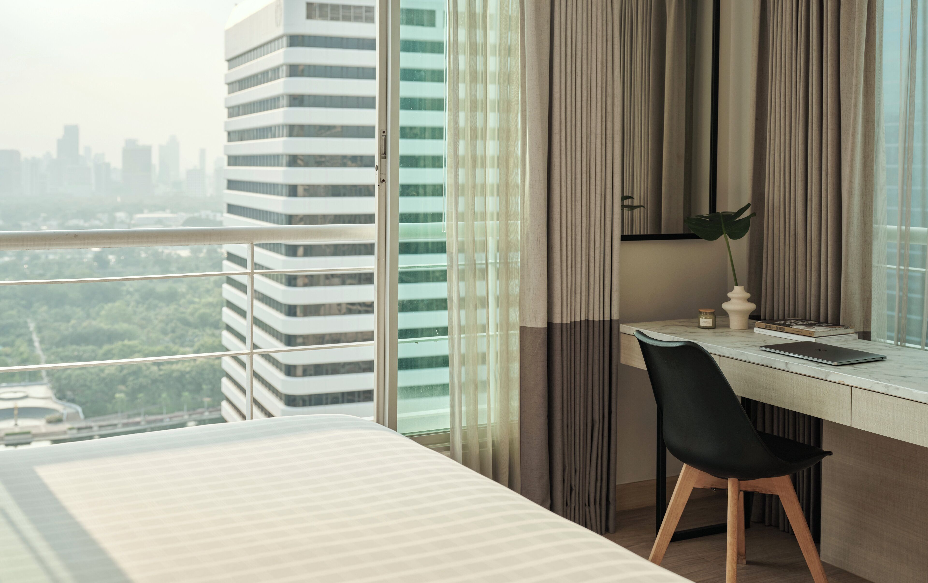 A modern hotel room with a large window, a desk with a laptop, a black chair, and a bed.