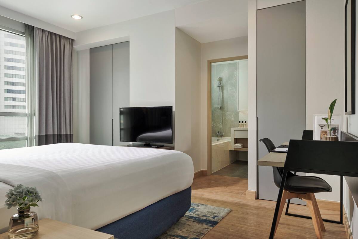 Une chambre d’hôtel moderne, dotée d’un grand lit, d’un bureau avec une chaise, d’une télévision et d’une salle de bain visible à travers une porte ouverte.