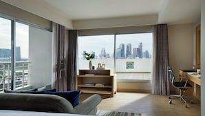 A modern hotel room with a large bed, a desk, a chair, a bookshelf, and a view of a city skyline through large windows.