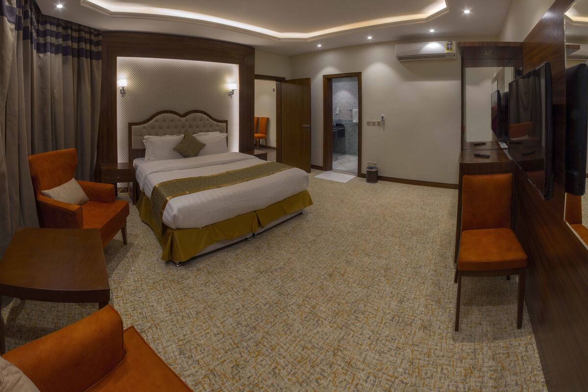 A hotel room with a large bed, two armchairs, a desk with a TV, and an air conditioning unit on the wall.