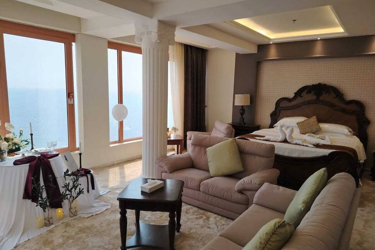 A hotel room with a bed, two sofas, a small table, and a view of the sea.
