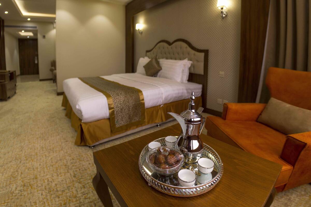 A hotel room with a bed, a sofa, a tray with a pitcher and cups, and a carpeted floor.