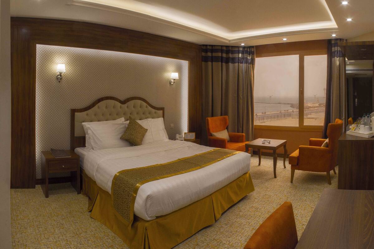 A hotel room with a large bed, two armchairs, a small table, and a view of the city.