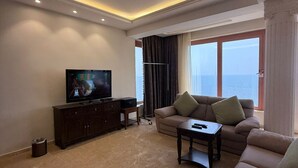 A modern living room with a flat-screen TV, dark wooden furniture, a beige sofa, and a view of the sea through large windows. - Al-Borg Al-Watheer Serviced Apartments (Jizan)