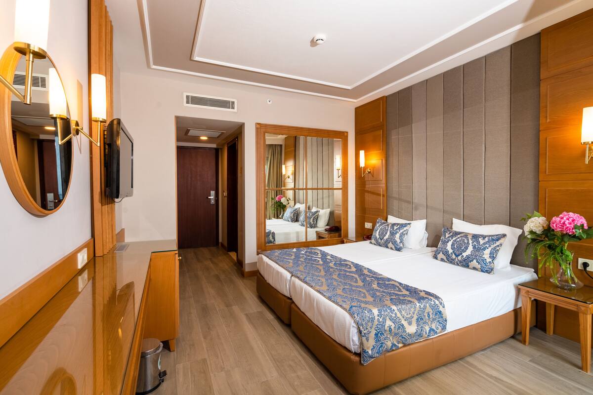 Habitación de hotel con una cama grande, un aparador de madera, un espejo y una mesita de noche con un jarrón de flores.