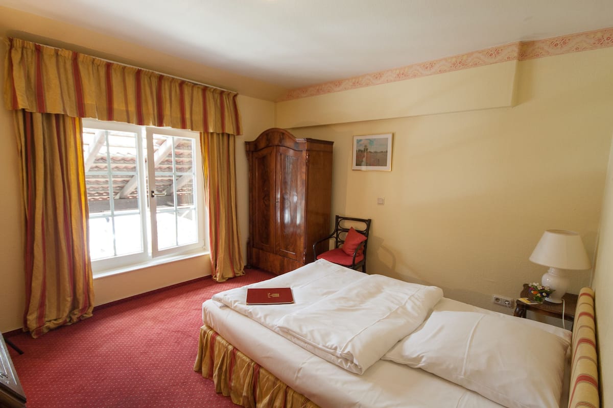 A hotel room with a bed, a wardrobe, a chair, a window with curtains, and a lamp on a bedside table.