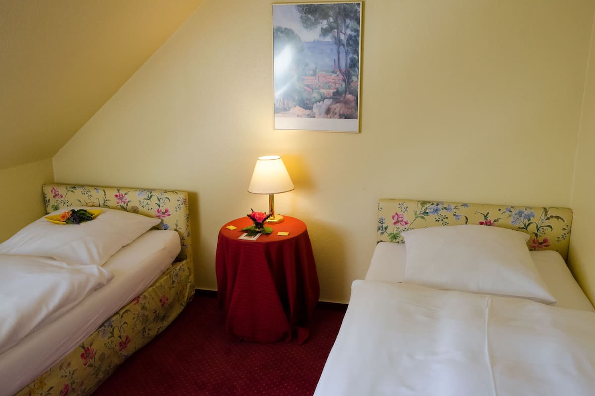 A room with two beds, a round table with a lamp, and a painting on the wall.
