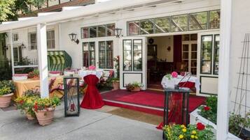 A hotel entrance with a red carpet, white walls, and green-framed windows. There are tables set up outside with red and white tablecloths, and potted plants with yellow and red flowers.