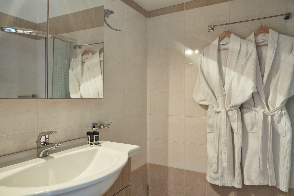 A modern bathroom with a large sink, a glass shower enclosure, and two white bathrobes hanging on a rack.