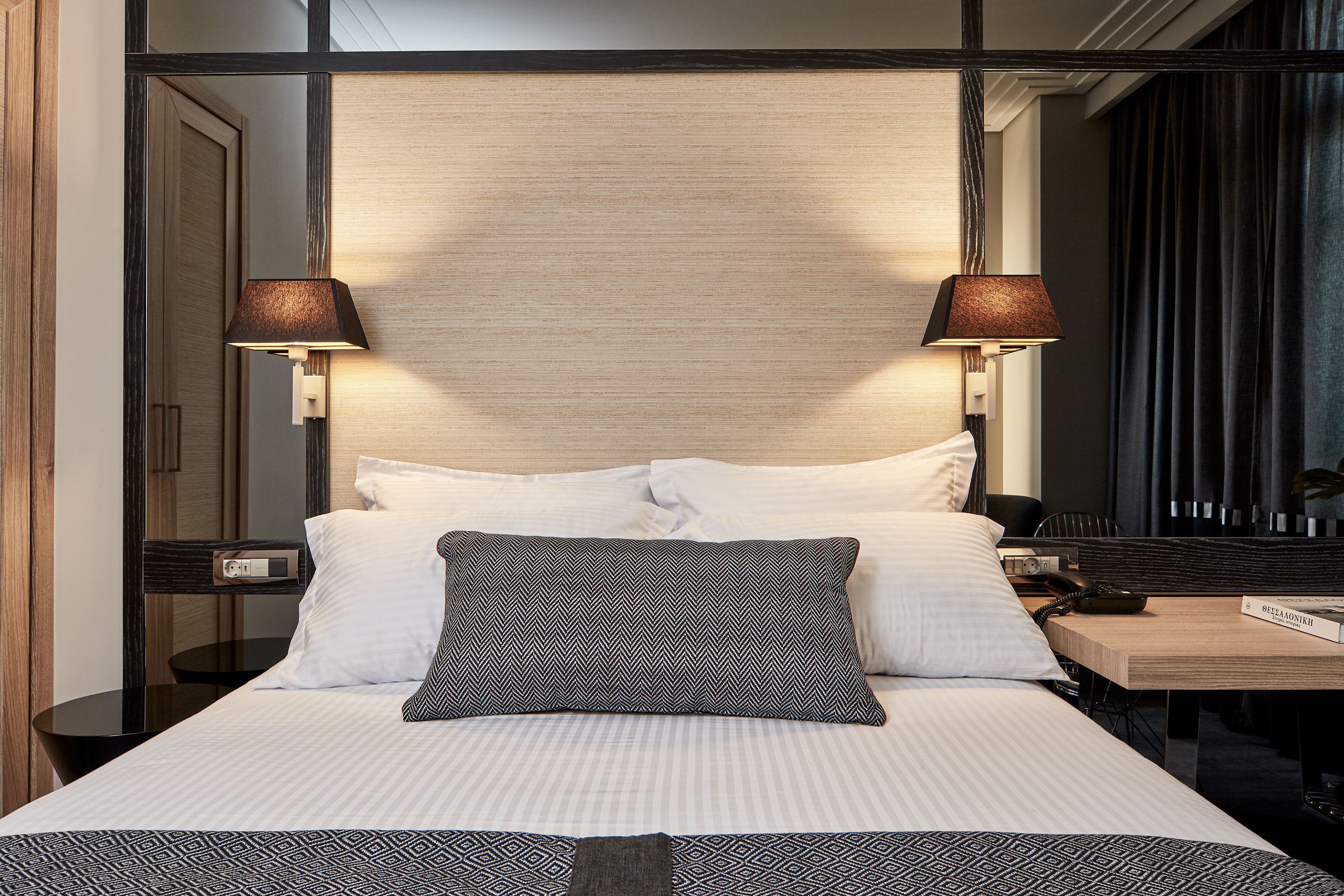 Una cama bien hecha con almohadas blancas y estampadas, una mesita de noche con lámpara y un espejo sobre la cama.