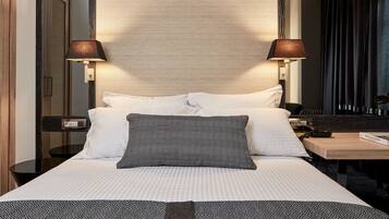 Una cama bien hecha con almohadas blancas y estampadas, una mesita de noche con lámpara y un espejo sobre la cama.