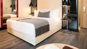 Una habitación de hotel moderna con una cama grande, una mesita de noche con una lámpara, una mesa redonda con un libro y vasos, y un espejo en la pared.