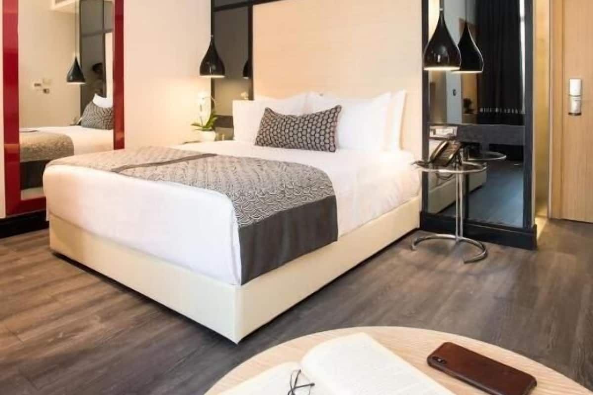 Une chambre d’hôtel moderne avec un grand lit, une table de chevet avec une lampe, une table ronde avec un livre et des lunettes, et un miroir au mur.