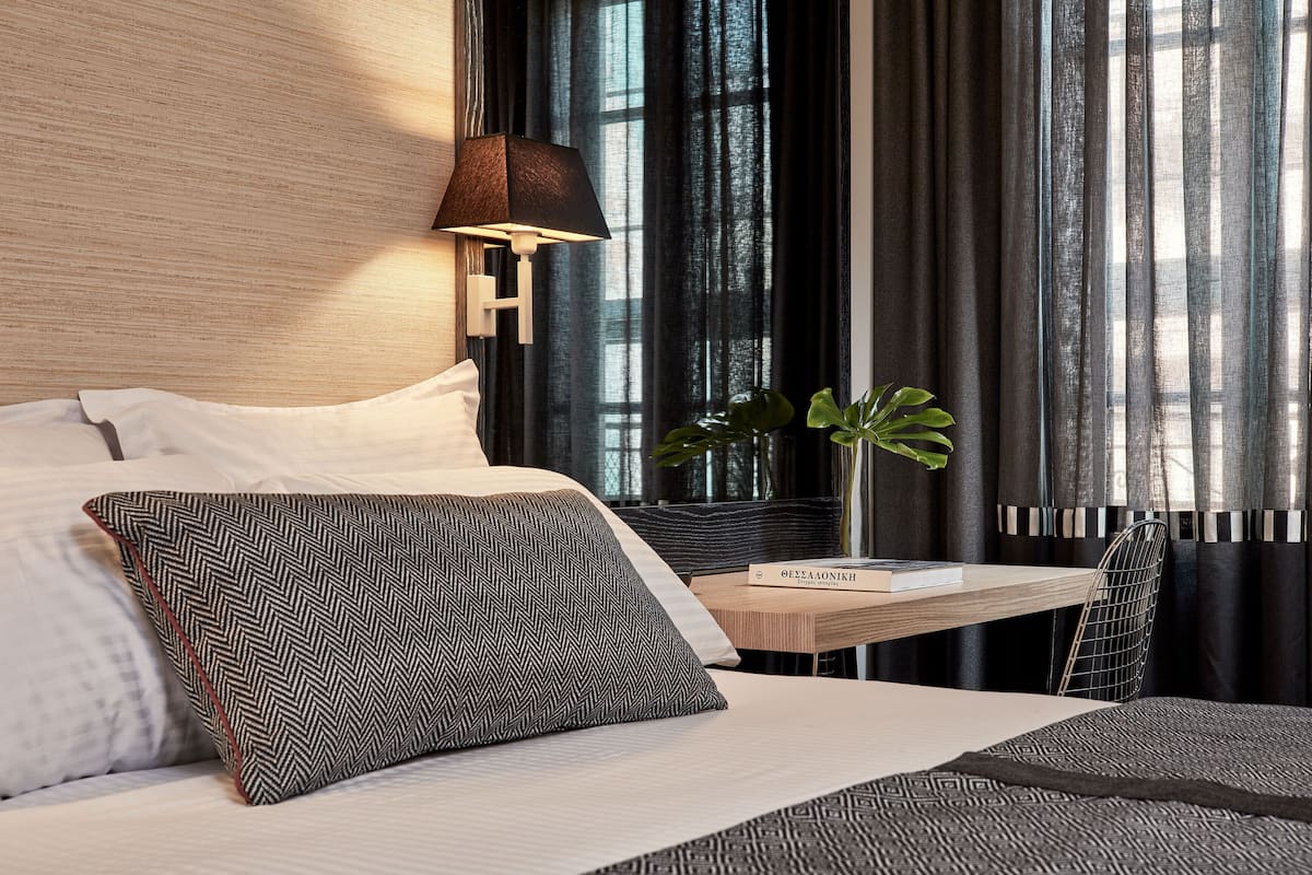 Quarto de hotel moderno com cama, mesa de cabeceira com um livro, abajur e uma planta.