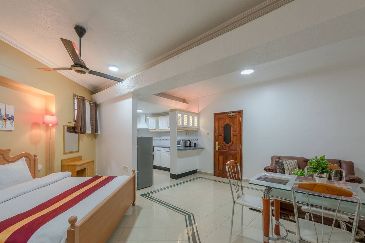 Ein Hotelzimmer mit Bett, Esstisch mit Stühlen, Deckenventilator und einer Küchenzeile im Hintergrund.