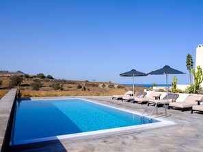 A rectangular swimming pool with a clear blue water, surrounded by lounge chairs and umbrellas, with a view of the sea and hills in the background. - Home of Lilies (Santorini)