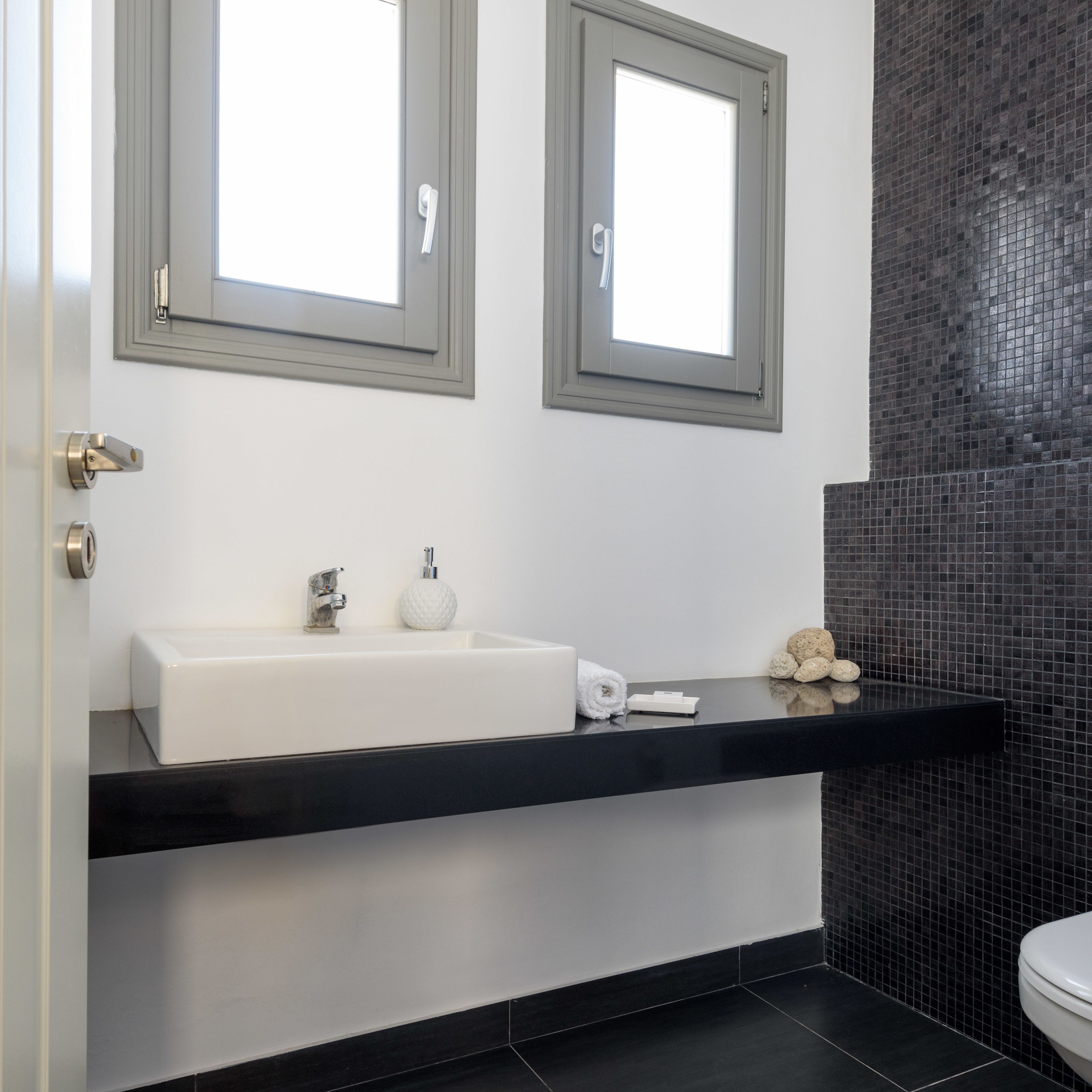 A modern bathroom with a white sink, two mirrors, a black countertop, and a black tiled wall.
