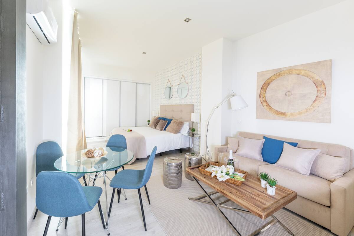 A modern hotel room with a glass dining table, blue chairs, a wooden coffee table, a beige sofa, and a large abstract painting.