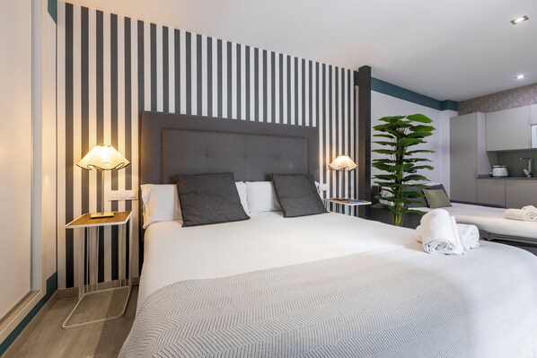 Une chambre d’hôtel avec un mur à rayures, un grand lit avec une tête de lit grise, deux lampes de chevet, une plante en pot et un miroir dans la salle de bain.
