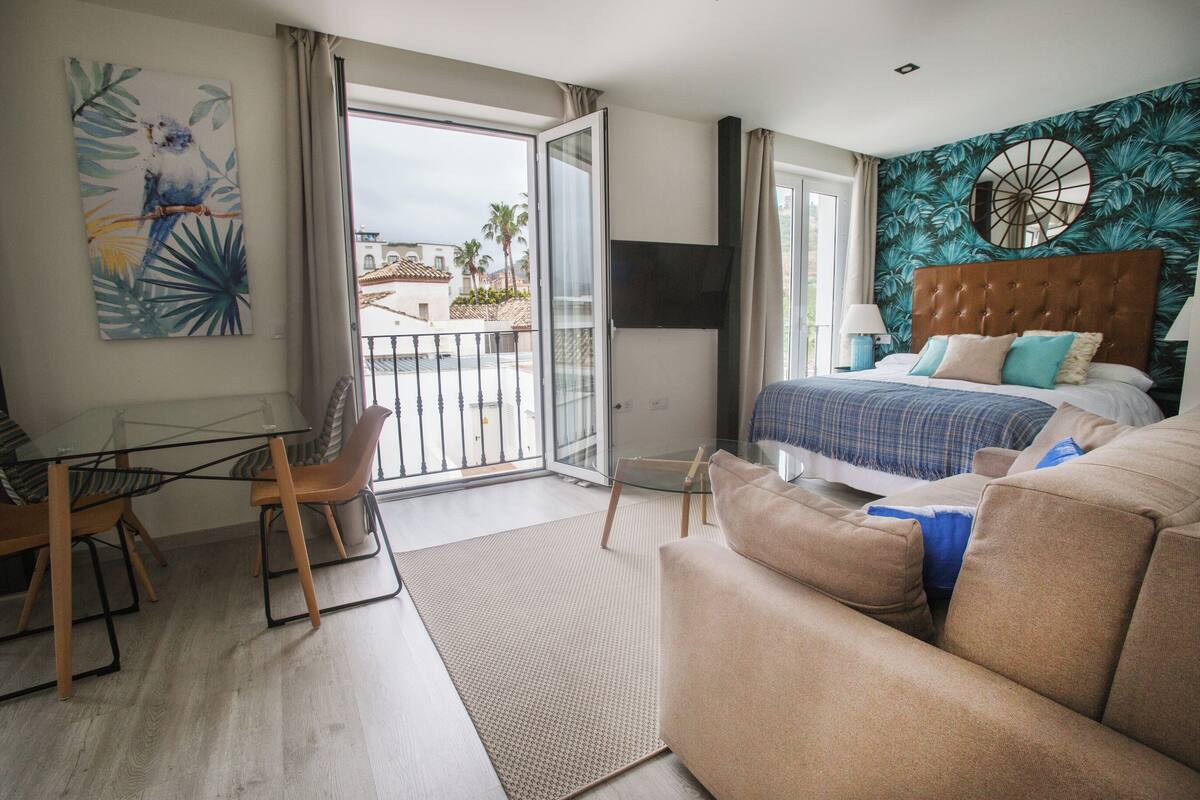 A hotel room with a bed, a sofa, a dining table with chairs, and a balcony with a view of palm trees and buildings.