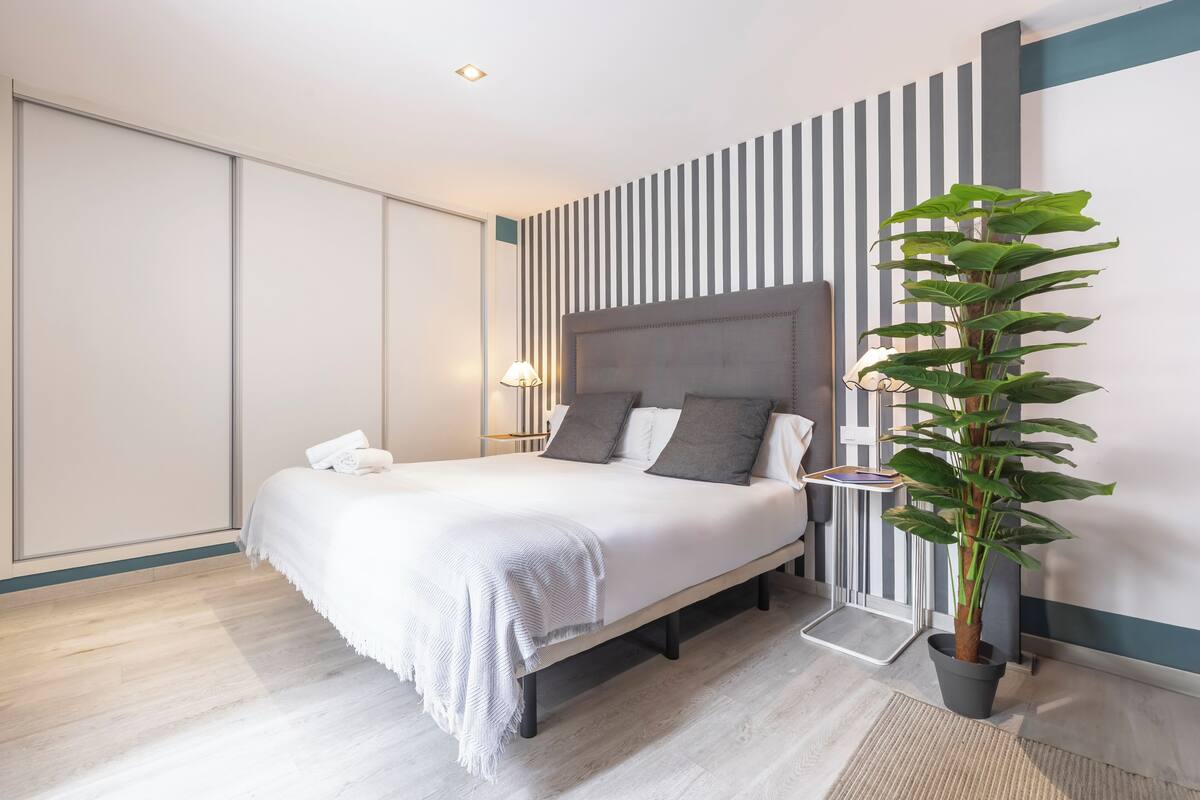 A modern bedroom with a bed, bedside table, lamp, and a potted plant.