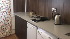 Kompakti keittiö, jossa on pesukone, vedenkeitin sekä pieni työtaso, jonka päällä on astioita ja ruokailuvälineitä.