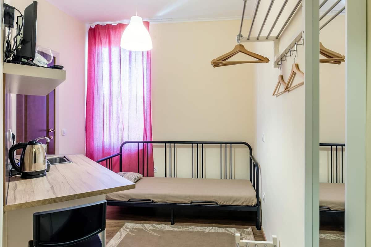 A compact room with a kitchenette, a single bed, a hanging light fixture, and a mirror.