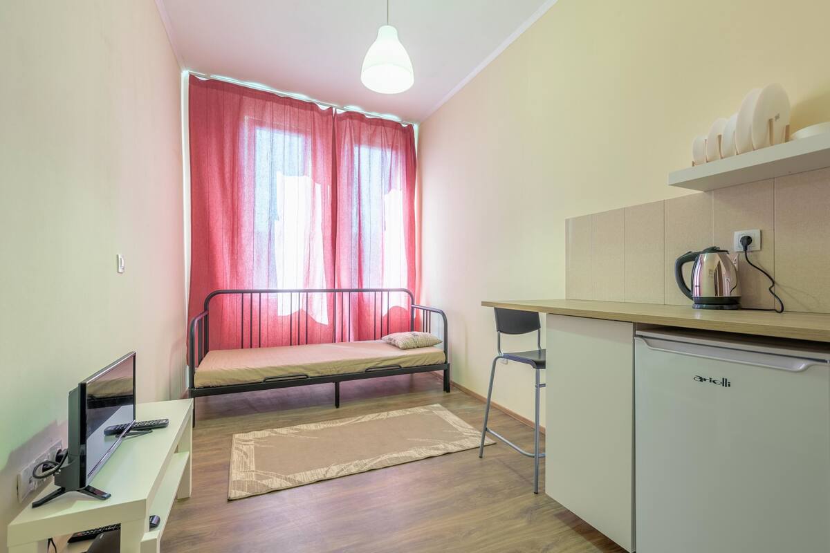 A compact room with a bed, a desk with a computer, a chair, a small kitchen area with a kettle, and a window with red curtains.