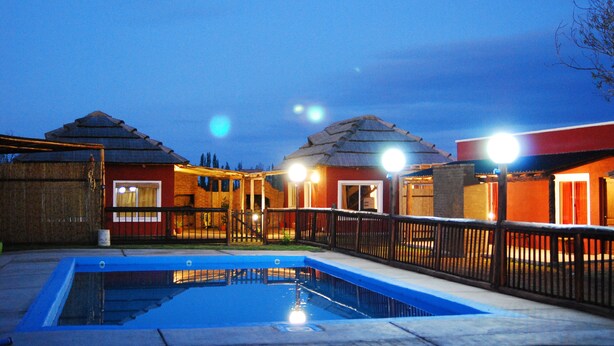 A swimming pool with a wooden deck and a fence, surrounded by buildings with red and orange walls and thatched roofs.