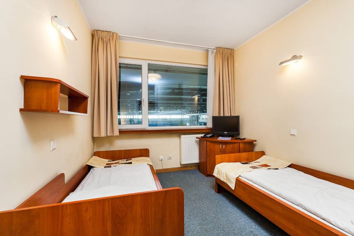 Une chambre d’hôtel avec deux lits en bois, une télévision, une petite étagère en bois et une fenêtre avec des rideaux.