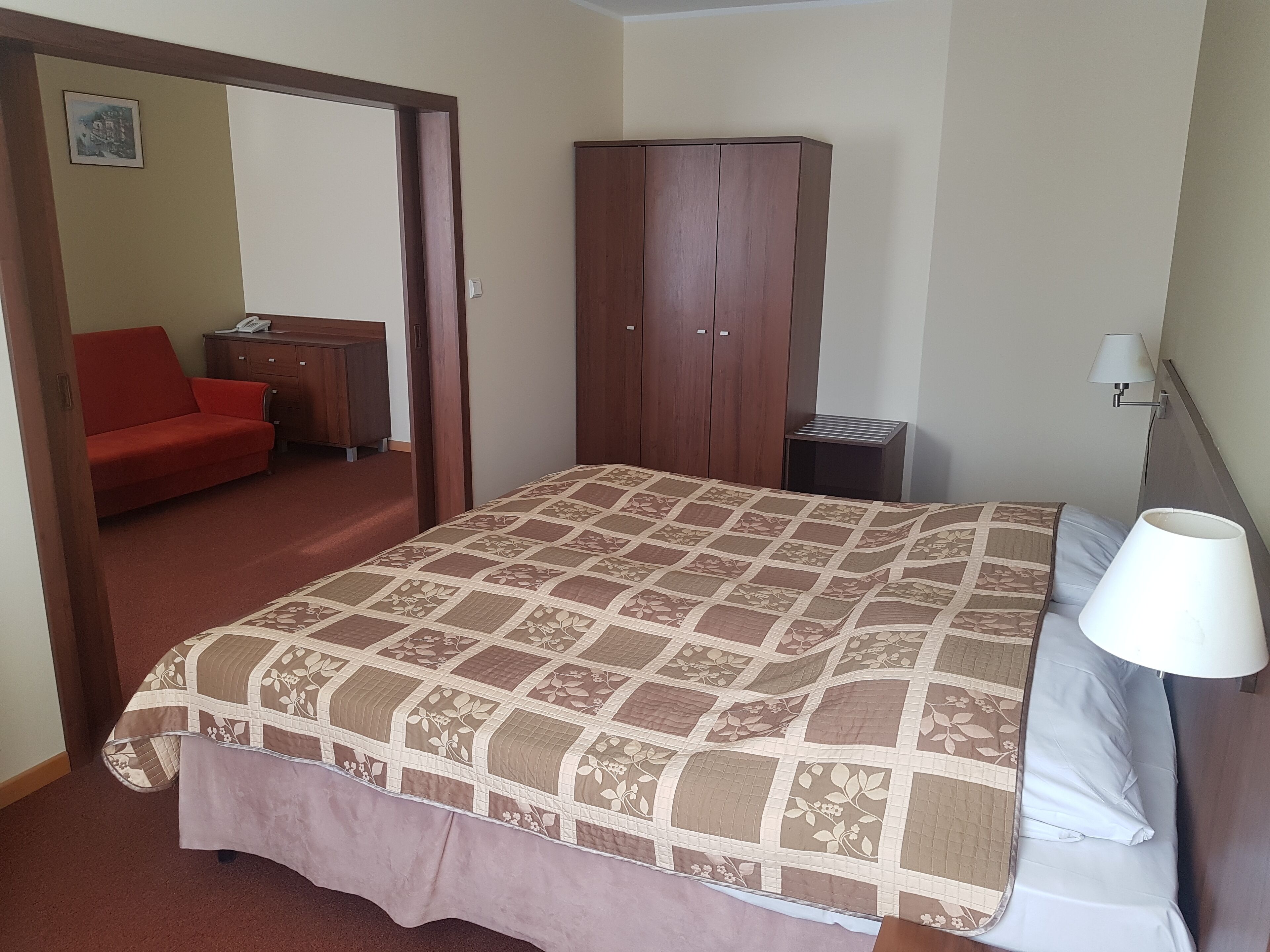 A bedroom with a bed, a wardrobe, a nightstand, a lamp, and a red sofa.