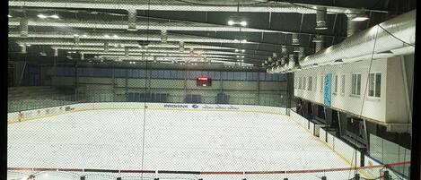 An indoor ice hockey rink with a netted enclosure, visible seating areas, and a scoreboard.