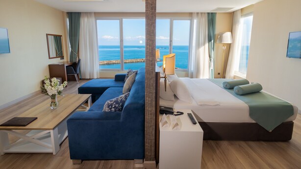 A hotel room with a large window offering a sea view, a bed, a sofa, a desk, and a lamp. - Tilya Resort Hotel (Akçaabat)