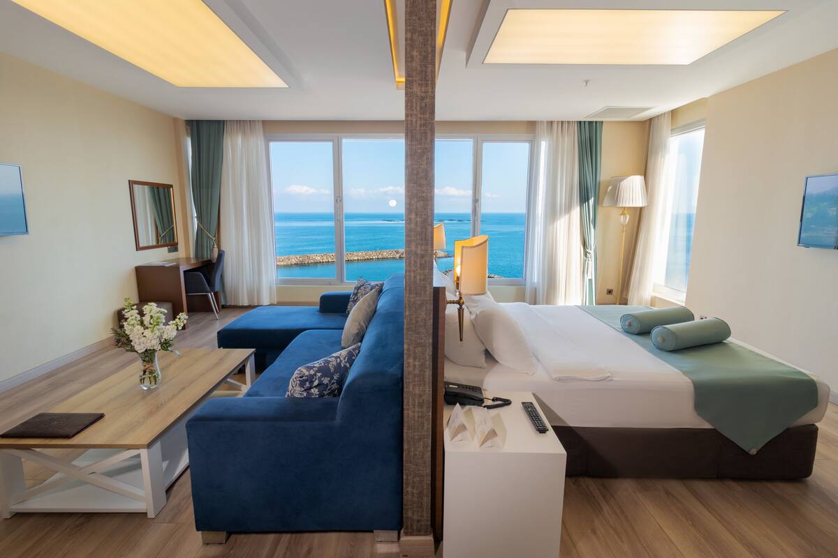 A hotel room with a large window offering a sea view, a bed, a sofa, a desk, and a lamp.