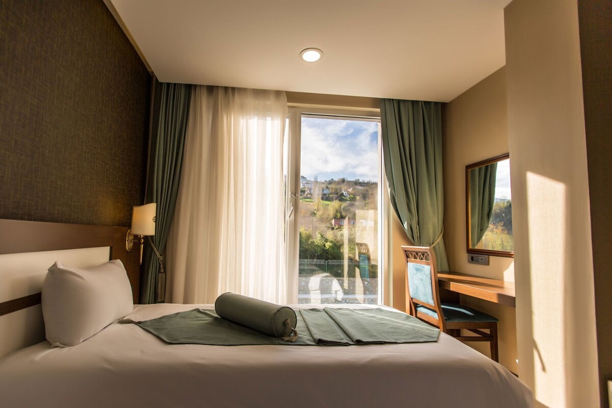 A hotel room with a bed, a desk, a chair, a lamp, and a view of the outside.