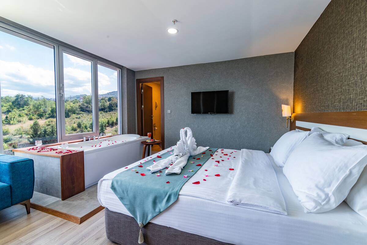 A hotel room with a large window, a bathtub, a bed with white linens, and a television on the wall.