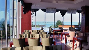 A dining area with a view of the sea, featuring a mix of wooden and upholstered chairs, a red accent wall, and a large window with curtains. - Tilya Resort Hotel (Akçaabat)