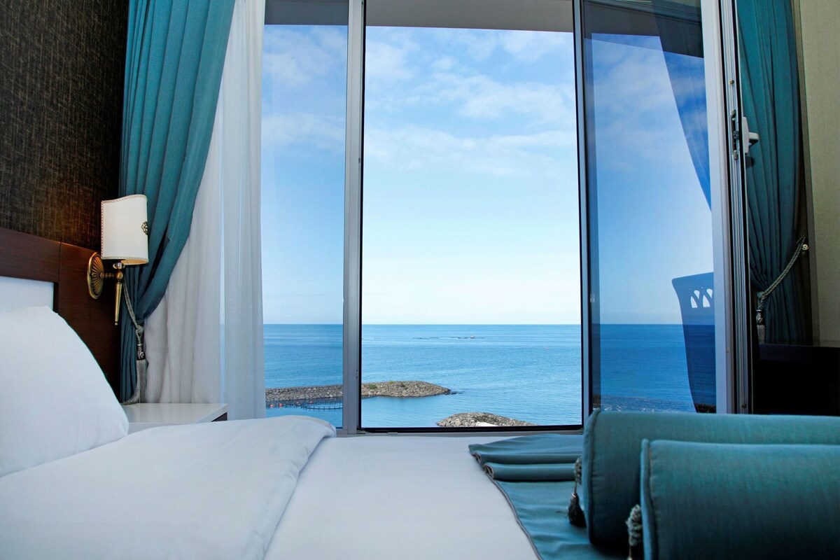 A hotel room with a bed, a nightstand, a lamp, a view of the sea through a glass door, and a chair.