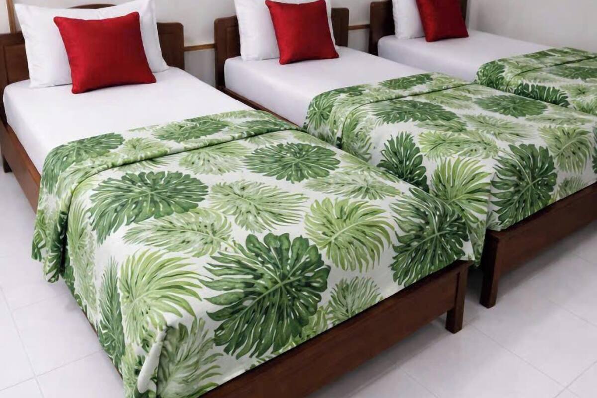 A hotel room with three single beds, each with white bedding and red pillows, and a large bed with a tropical leaf-patterned bedspread.