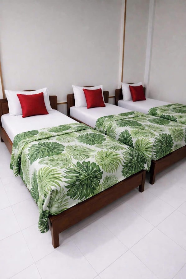 A hotel room with three single beds, each with white bedding and red pillows, and a large bed with a tropical leaf-patterned bedspread.