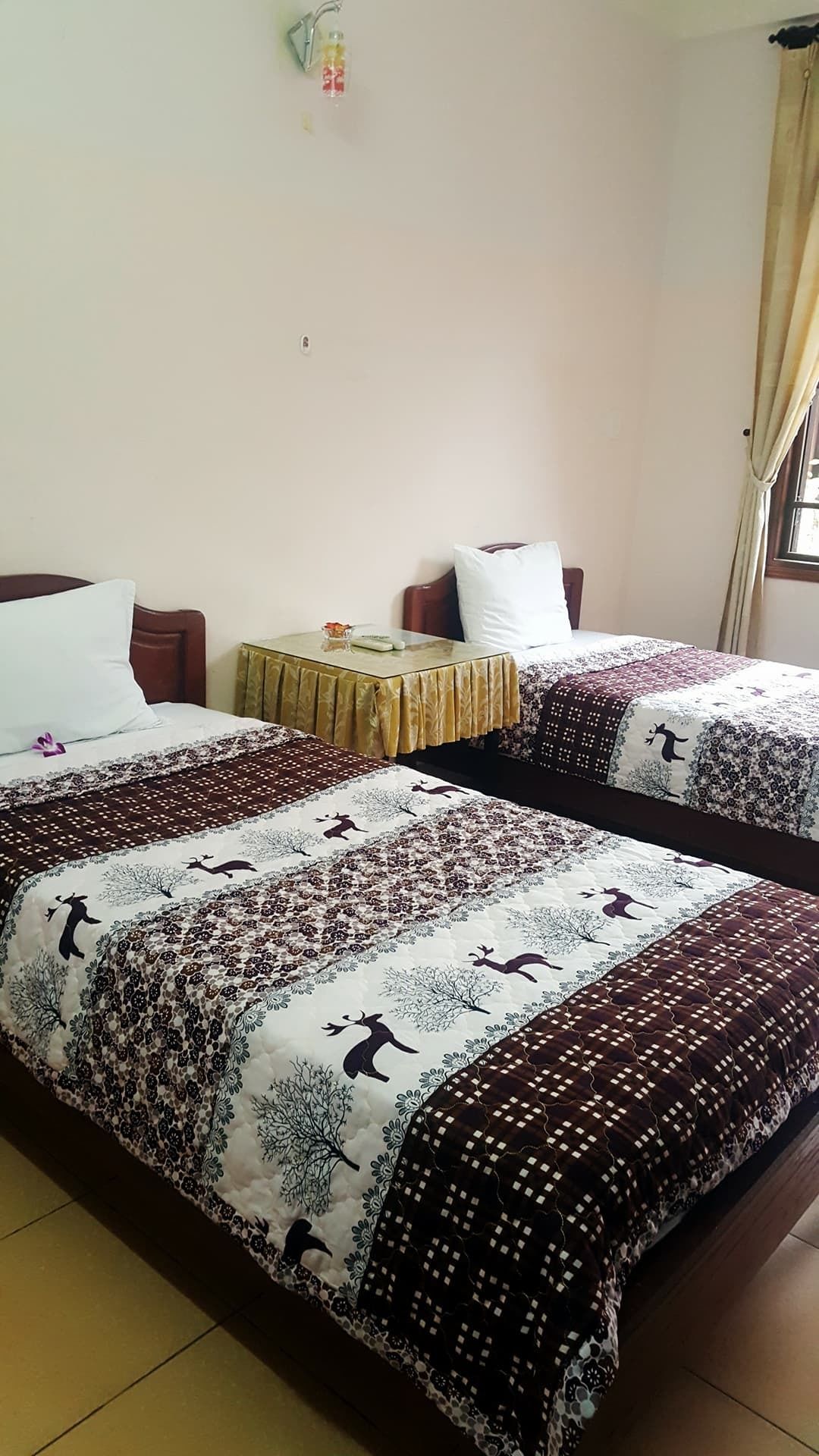 A hotel room with two beds, each with a patterned bedspread, a small table with a lamp, and a window with curtains.